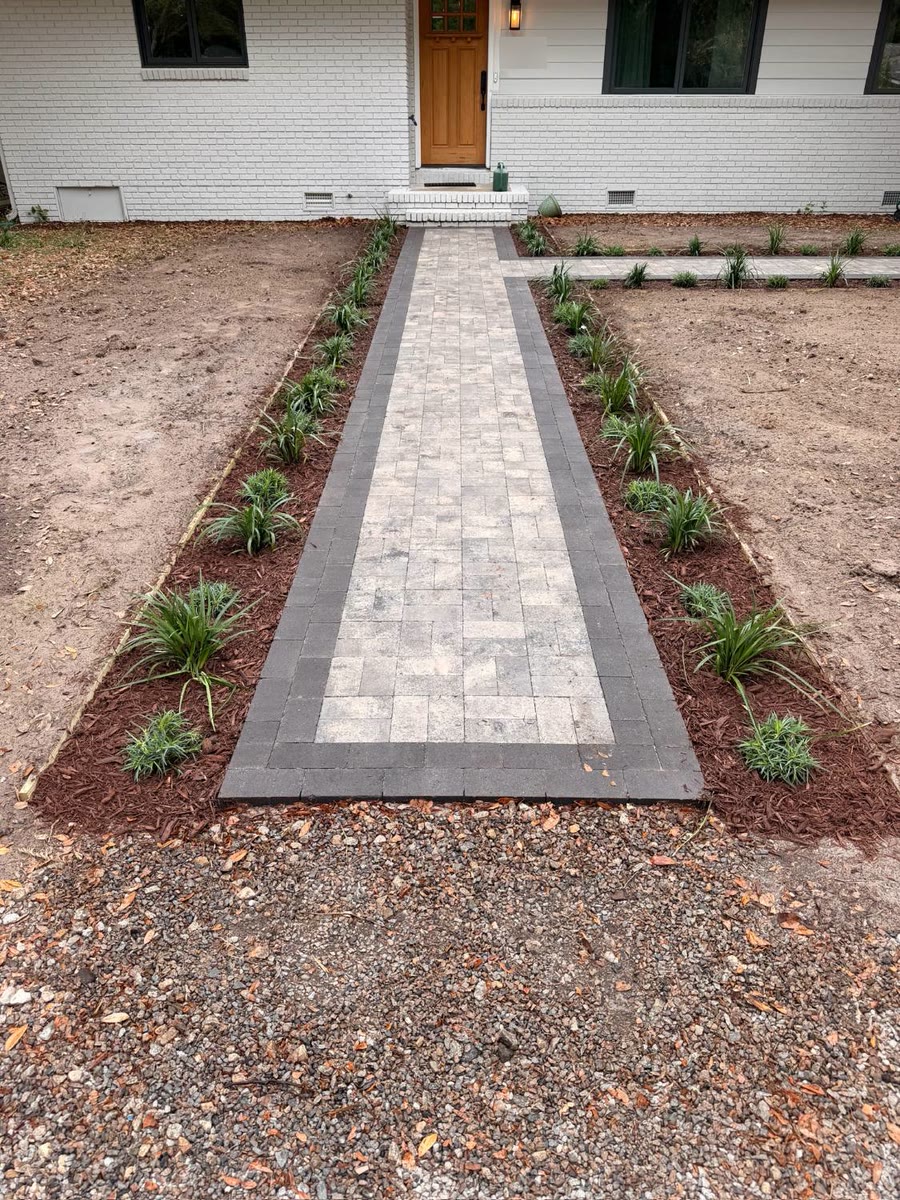Paver front walk with a charcoal border and newly planted, mulched beds on both sides, leading to a front door